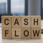 Une photographie en gros plan de huit blocs de bois carrés empilés sur deux rangées sur une table de bureau en marbre poli. Les blocs affichent les lettres noires "CASH" sur la rangée du haut et "FLOW" sur la rangée du bas représentant le mot "cash flow". En arrière-plan, un cadre de bureau moderne