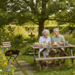 Une photographie lumineuse et naturelle d'un couple de retraités souriants, prenant le petit-déjeuner dans un jardin sauvage et fleuri en été. Symbolisant la sérénité après avoir obtenu un prêt viager hypothécaire.