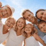 Famille multigénérationnelle souriante réunissant grands-parents, parents et enfants, photographiée en contre-plongée à l’extérieur sous un ciel bleu lumineux, ambiance chaleureuse et rassurante.