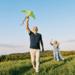 Un couple de seniors souriants profite d'une journée en plein air dans une prairie verdoyante ; l'homme court au premier plan en tenant un cerf-volant vert, tandis que la femme aux longs cheveux gris le salue joyeusement en arrière-plan. Ils profitent de la retraite,